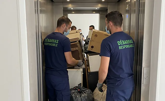Deux travailleurs de Débarras Responsable (uniforme bleu, texte vert) chargeant méticuleusement des sacs de déchets compactés et triés dans l’ascenseur d’un immeuble d’habitation moderne.