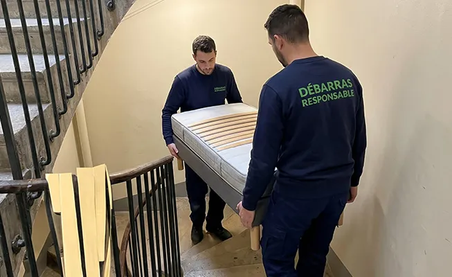 Deux professionnels Débarras Responsable descendant en synchro une lourde machine à coudre vintage en fonte dans un escalier colimaçon de grenier étroit.