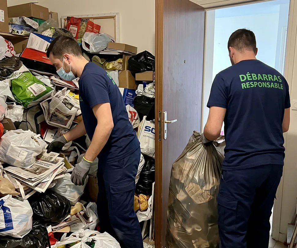 Deux professionnels de Débarras Responsable (uniforme bleu, texte vert) gèrent un amas de déchets et d’objets accumulés à l’entrée d’un appartement saturé (Syndrome de Diogène).
