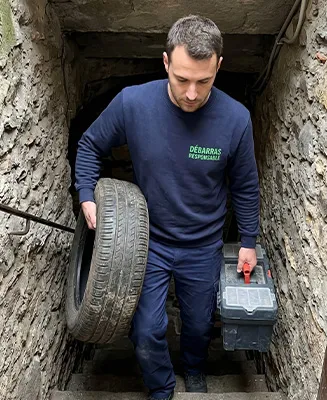 Professionnel de Débarras Responsable manœuvrant un vieux pneu et une boîte à outils lourde avec technique dans un escalier de cave en pierre très étroit.