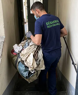 Professionnel de Débarras Responsable (uniforme bleu, texte vert) manœuvrant délicatement un gros volume de sacs de déchets lourds avec technique dans un couloir d’appartement étroit.