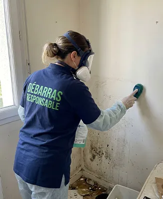 Travailleuse de Débarras Responsable (uniforme bleu, texte vert) effectuant un nettoyage désinfectant approfondi d’un mur de cuisine couvert de grime dans un logement insalubre.